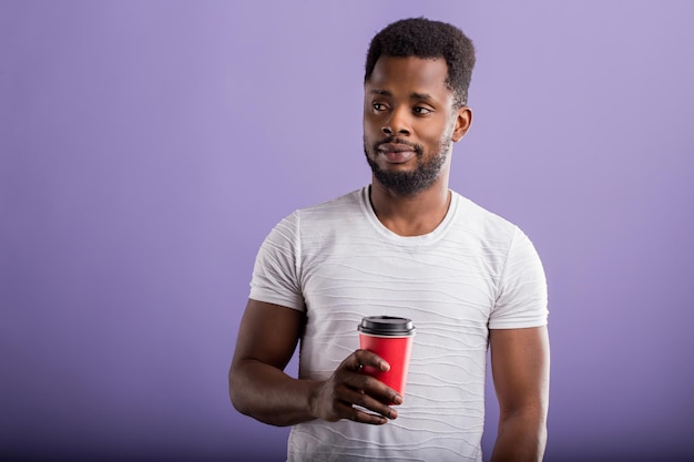 Young african man with short curly hair, enjoying free time, wearing casual white tshirt, holding takeaway coffee, isolated over purple background, drinking tasty beverage. People and leisure.