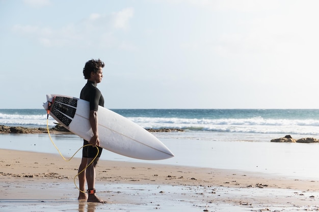 Young african surfer standing on the beach shore watching the waves