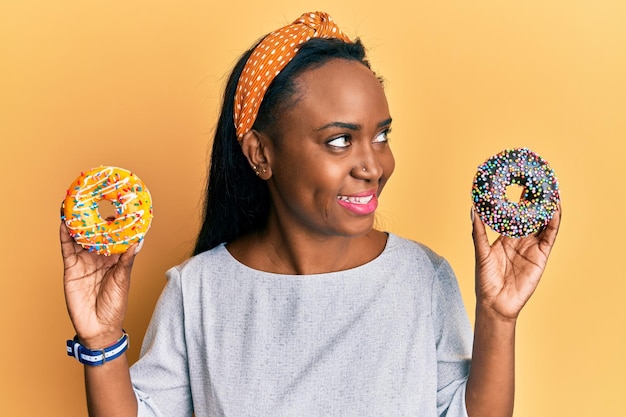 Young african woman holding tasty colorful doughnuts smiling looking to the side and staring away thinking