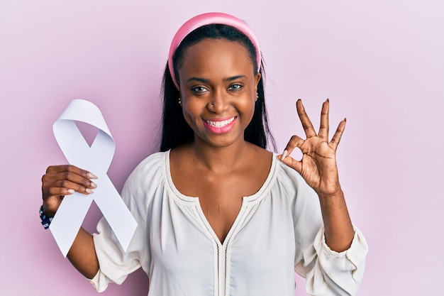 Young african woman holding white ribbon doing ok sign with fingers, smiling friendly gesturing excellent symbol