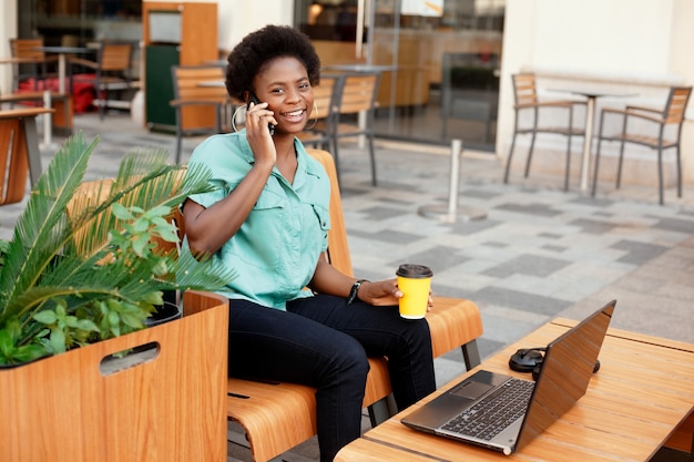 A young African woman is sitting in a street cafe, drinking coffee, talking on the phone and working on a laptop.