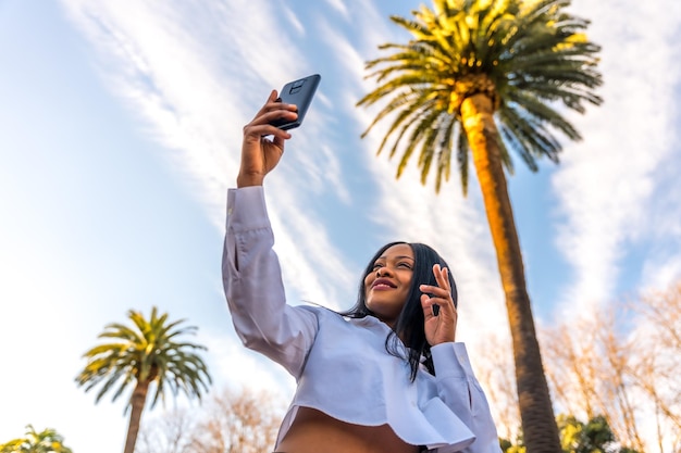 Young african woman posing in white clothes in a tropical place with palm trees at sunset enjoying the summer taking a selfie