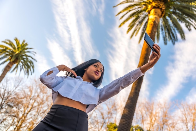 Young african woman posing in white clothes in a tropical place with palm trees at sunset taking a selfie