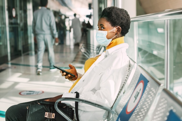 A young African woman in a protective mask sits in a chair in the waiting room and reads emails on her phone. Social distance, pandemic.