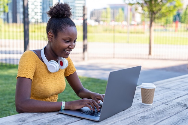 Young african woman using laptop outdoor blogger creating content in the city while drinking takeaway coffee smart work concept possibility to relocate activities