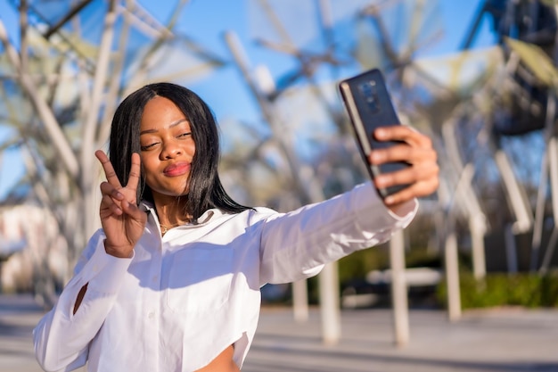 Young african woman in white clothes in the city at sunset video call making a victory gesture