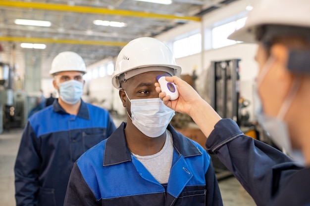 Young African worker in workwear and protective mask having his body temperature measured by electronic thermometer held by colleague