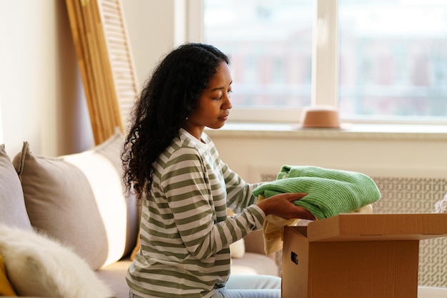 Young africanamerican female packing stuff in cardboard box moving out and giving clothes away