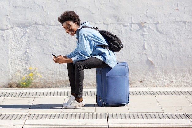 Photo young afro american woman sitting on suitcase at railway platform with mobile phone