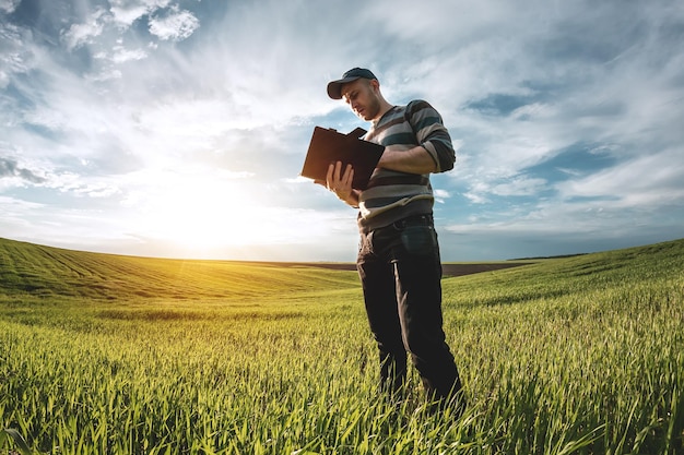 A young agronomist holds a folder in his hands on a green wheat field. A farmer makes notes on the background of agricultural land during sunset. Man in a cap with a folder of documents