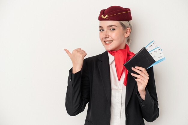 Young air hostess caucasian woman holding passport isolated on white background points with thumb finger away laughing and carefree