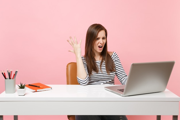 Young angry irritated woman spreading hand working on project while sitting at office with laptop