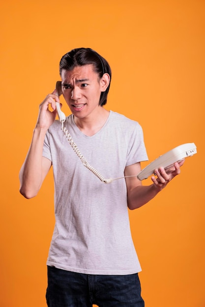 Young angry man talking on landline phone. Irritated asian teenager shouting, holding retro telephone, person with annoyed facial expression having conversation, answering call