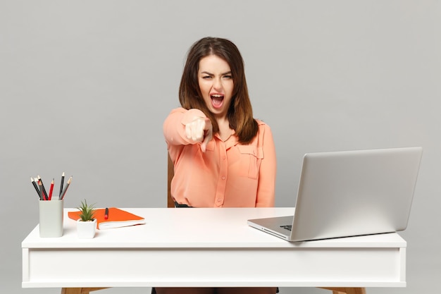 Young angry woman in pastel clothes swearing pointing index finger on camera sit work at white desk with pc laptop isolated on gray background. Achievement business career concept. Mock up copy space.