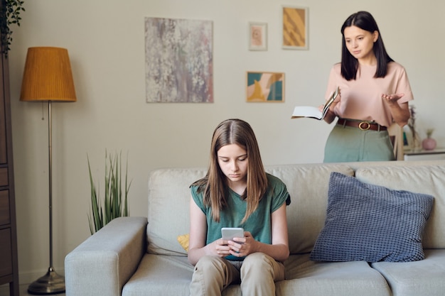 Young annoyed brunette mother with copybook of her teenage daughter standing by couch behind the girl texting in mobile phone