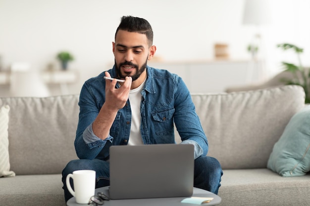 Young Arab guy using laptop computer and talking on mobile phone working remotely having business meeting at home