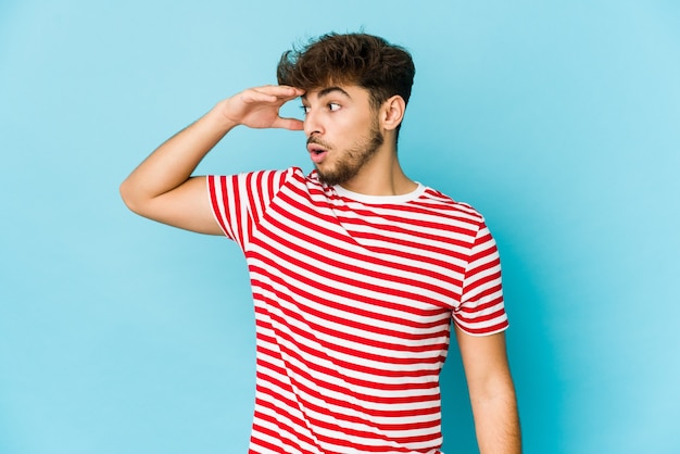 Young arab man on blue looking far away keeping hand on forehead.