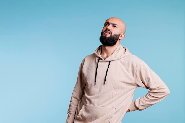 Young arab man holding hand on lower back while having spine problems. Bearded person looking upwards in pain and grimacing while suffering from backache and sore muscles cramps