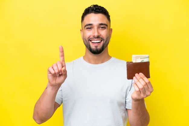 Young Arab man holding a wallet isolated on yellow background pointing up a great idea