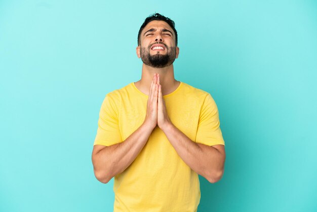 Young arab man isolated on blue background keeps palm together. Person asks for something