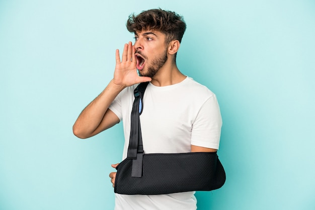 Young arab man with broken hand isolated on blue background shouting and holding palm near opened mouth.