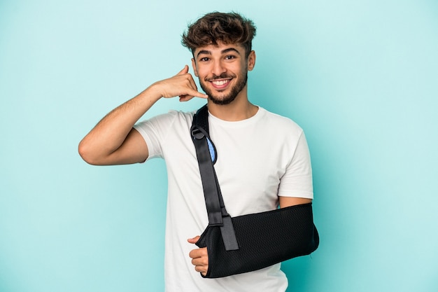 Young arab man with broken hand isolated on blue background showing a mobile phone call gesture with fingers.