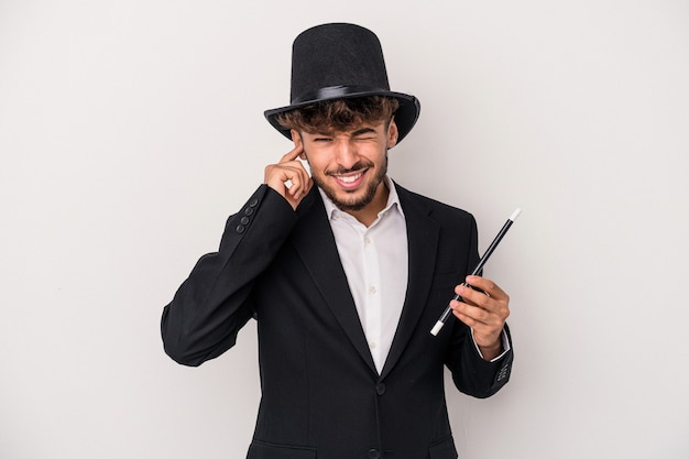 Young arab wizard man holding a wand isolated on white background covering ears with hands.