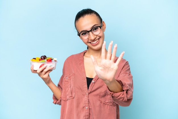 Young Arab woman holding a bowl of fruit isolated on blue background counting five with fingers