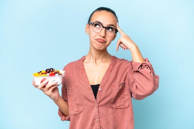 Young Arab woman holding a bowl of fruit isolated on blue background having doubts and thinking