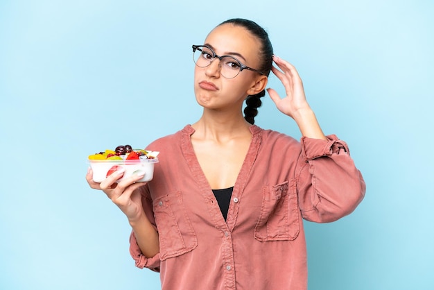 Young Arab woman holding a bowl of fruit isolated on blue background having doubts