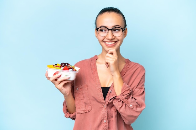 Young Arab woman holding a bowl of fruit isolated on blue background looking to the side and smiling