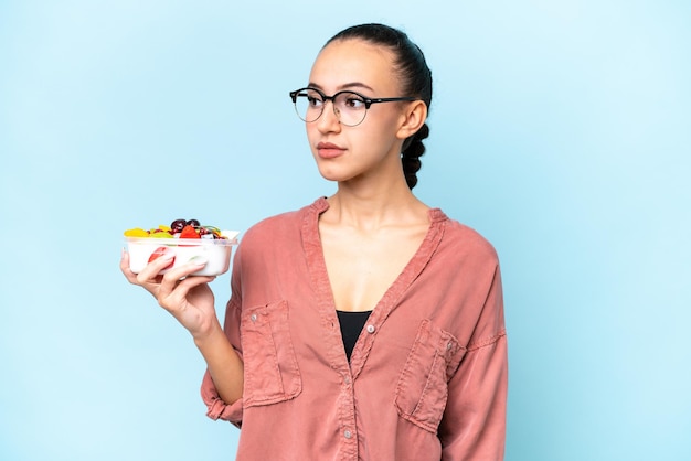 Young Arab woman holding a bowl of fruit isolated on blue background looking to the side