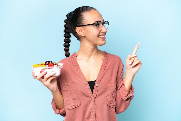 Young Arab woman holding a bowl of fruit isolated on blue background pointing up a great idea