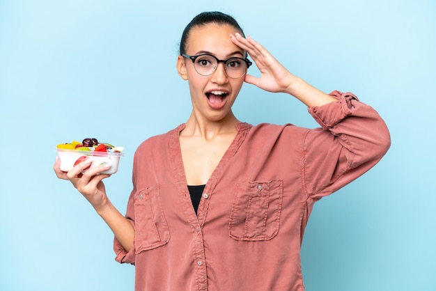 Young Arab woman holding a bowl of fruit isolated on blue background with surprise expression