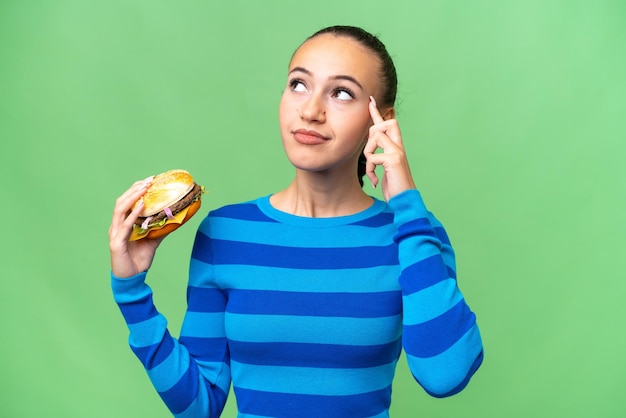 Young Arab woman holding a burger over isolated background having doubts and thinking