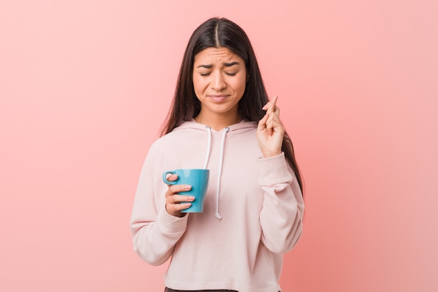 Young arab woman holding a cup crossing fingers for having luck