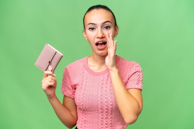 Young Arab woman holding a wallet over isolated background shouting with mouth wide open