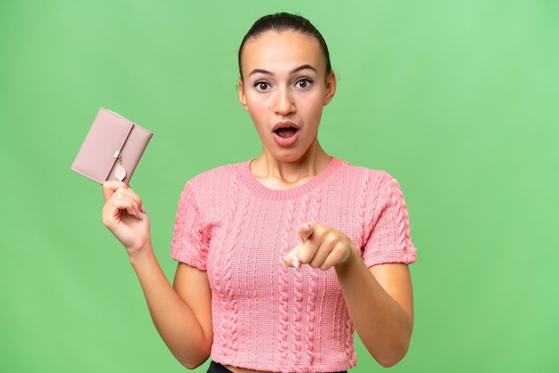 Young Arab woman holding a wallet over isolated background surprised and pointing front