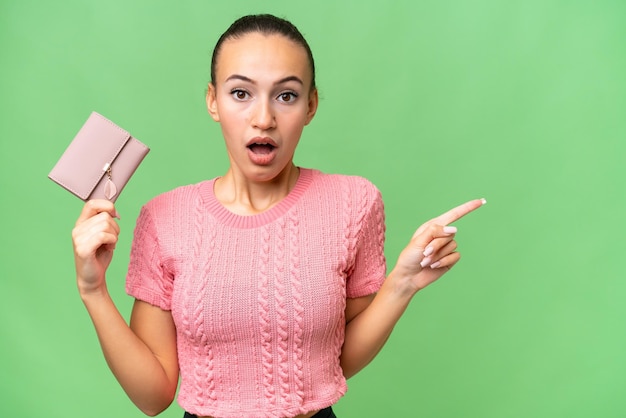 Young Arab woman holding a wallet over isolated background surprised and pointing side