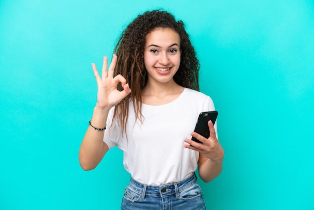 Young Arab woman isolated on blue background using mobile phone and doing OK sign