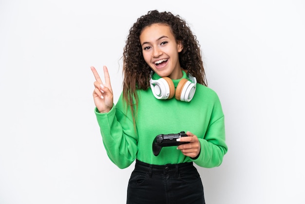 Young Arab woman playing with a video game controller isolated on white background smiling and showing victory sign