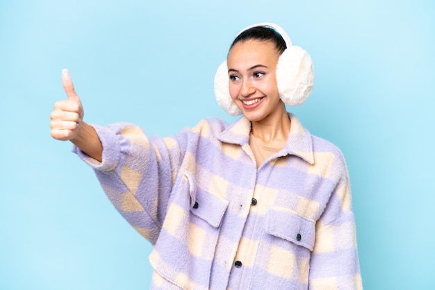 Young Arab woman wearing winter muffs isolated on blue background giving a thumbs up gesture