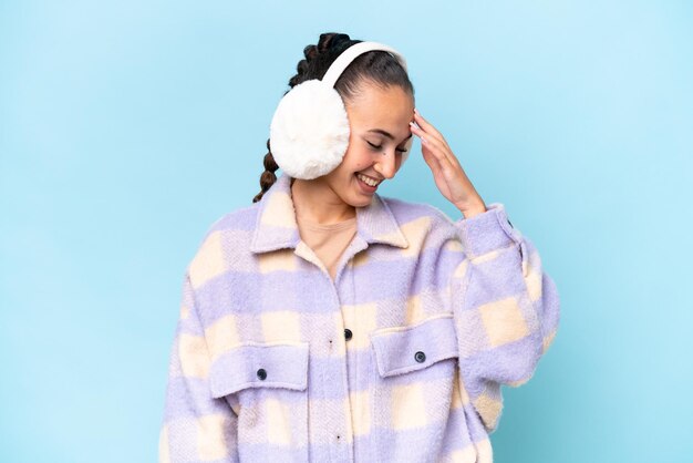 Young Arab woman wearing winter muffs isolated on blue background laughing