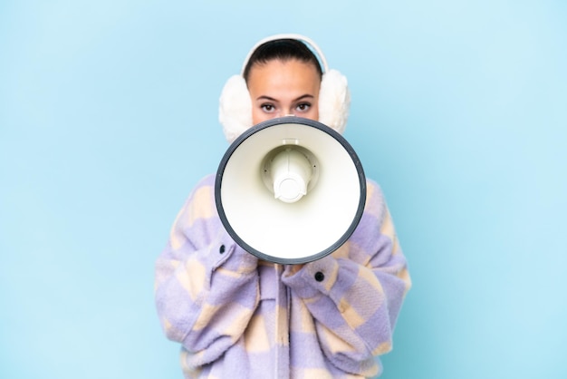 Young Arab woman wearing winter muffs isolated on blue background shouting through a megaphone