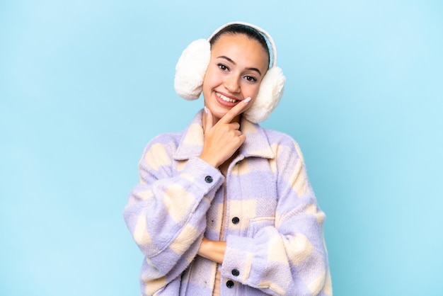 Young Arab woman wearing winter muffs isolated on blue background smiling