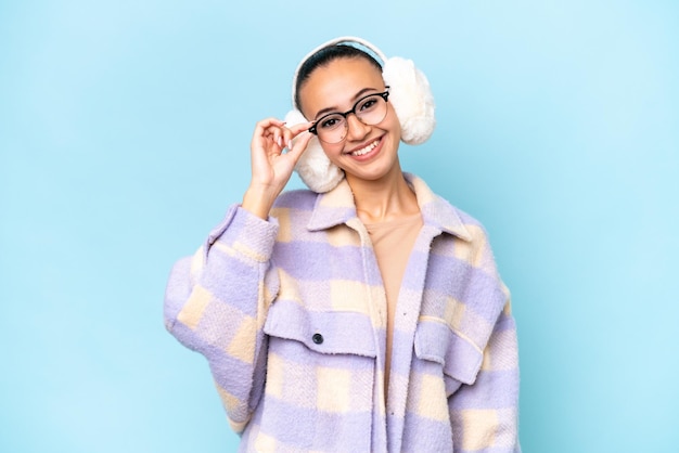 Young Arab woman wearing winter muffs isolated on blue background with glasses and happy