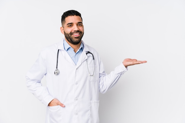 Young arabian doctor man isolated showing a copy space on a palm and holding another hand on waist.