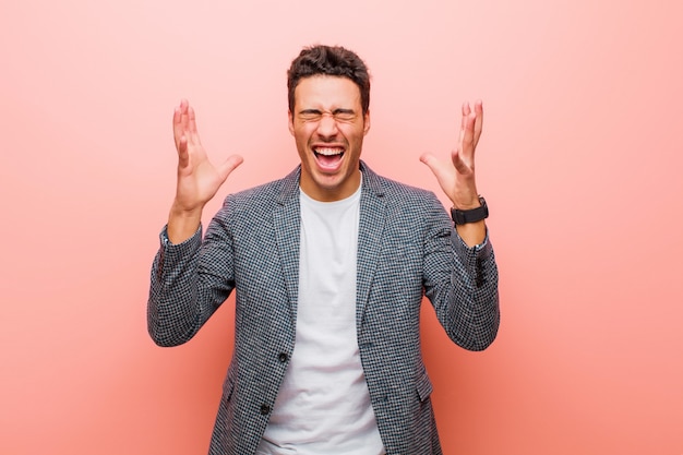 Young arabian man furiously screaming, feeling stressed and annoyed with hands up in the air saying why me against pink wall