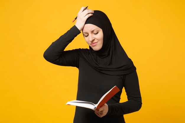 Young arabian muslim woman in hijab black clothes hold notebook, writing, prepare to exam isolated on yellow wall  portrait. People religious lifestyle concept. .