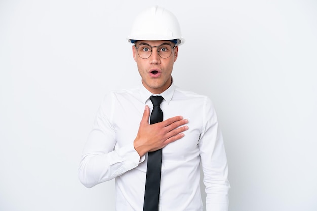 Young architect caucasian man with helmet and holding blueprints isolated on white background surprised and shocked while looking right